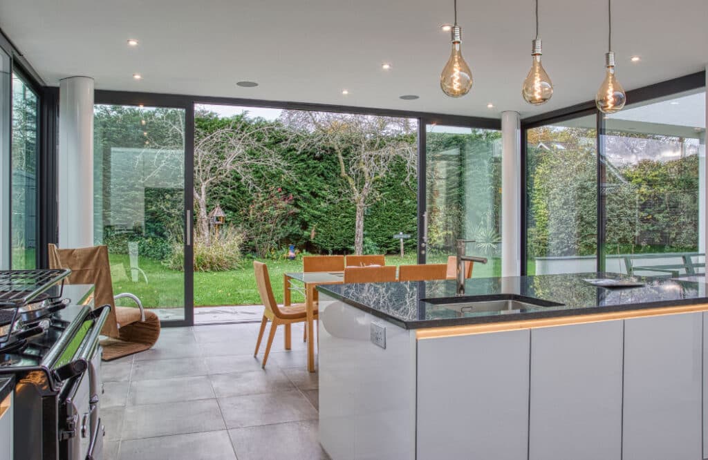 Modern kitchen and dining area with glass walls overlooking a garden. Features an island with a stovetop, wooden dining table with chairs, pendant lights, and tiled flooring.