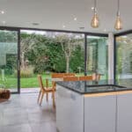 Thumbnail of http://Modern%20kitchen%20and%20dining%20area%20with%20glass%20walls%20overlooking%20a%20garden.%20Features%20an%20island%20with%20a%20stovetop,%20wooden%20dining%20table%20with%20chairs,%20pendant%20lights,%20and%20tiled%20flooring.