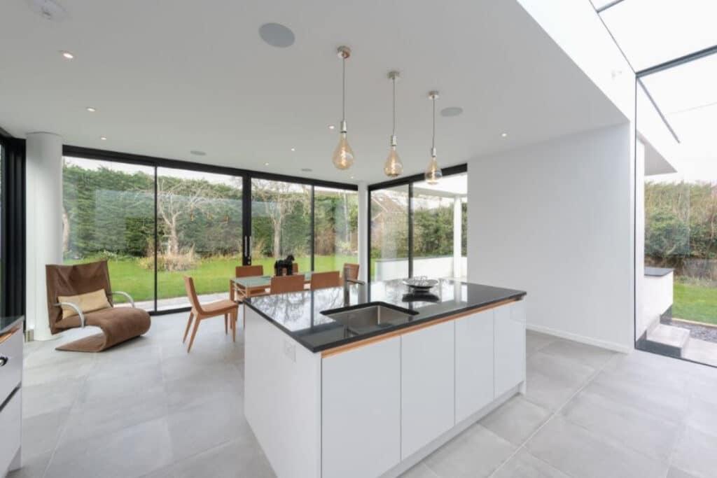 Modern open-plan kitchen and dining area with white cabinets, a black countertop island, wooden dining table, pendant lights, glass walls, and a view of a garden.