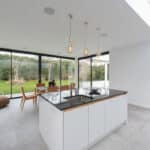 Thumbnail of http://Modern%20open-plan%20kitchen%20and%20dining%20area%20with%20white%20cabinets,%20a%20black%20countertop%20island,%20wooden%20dining%20table,%20pendant%20lights,%20glass%20walls,%20and%20a%20view%20of%20a%20garden.