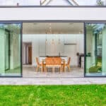 Thumbnail of http://Modern%20dining%20area%20with%20a%20table%20and%20chairs%20viewed%20from%20outside%20through%20large%20glass%20sliding%20doors.%20Green%20lawn%20in%20the%20foreground%20and%20white%20minimalist%20interior.