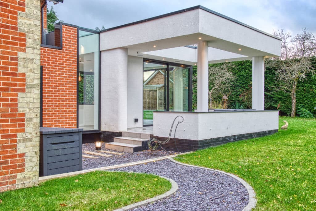 Modern building extension with large glass windows and flat roof, connected to a brick house. Curved gravel path leads through a green lawn, with trees and hedges in the background.