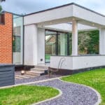 Thumbnail of http://Modern%20building%20extension%20with%20large%20glass%20windows%20and%20flat%20roof,%20connected%20to%20a%20brick%20house.%20Curved%20gravel%20path%20leads%20through%20a%20green%20lawn,%20with%20trees%20and%20hedges%20in%20the%20background.