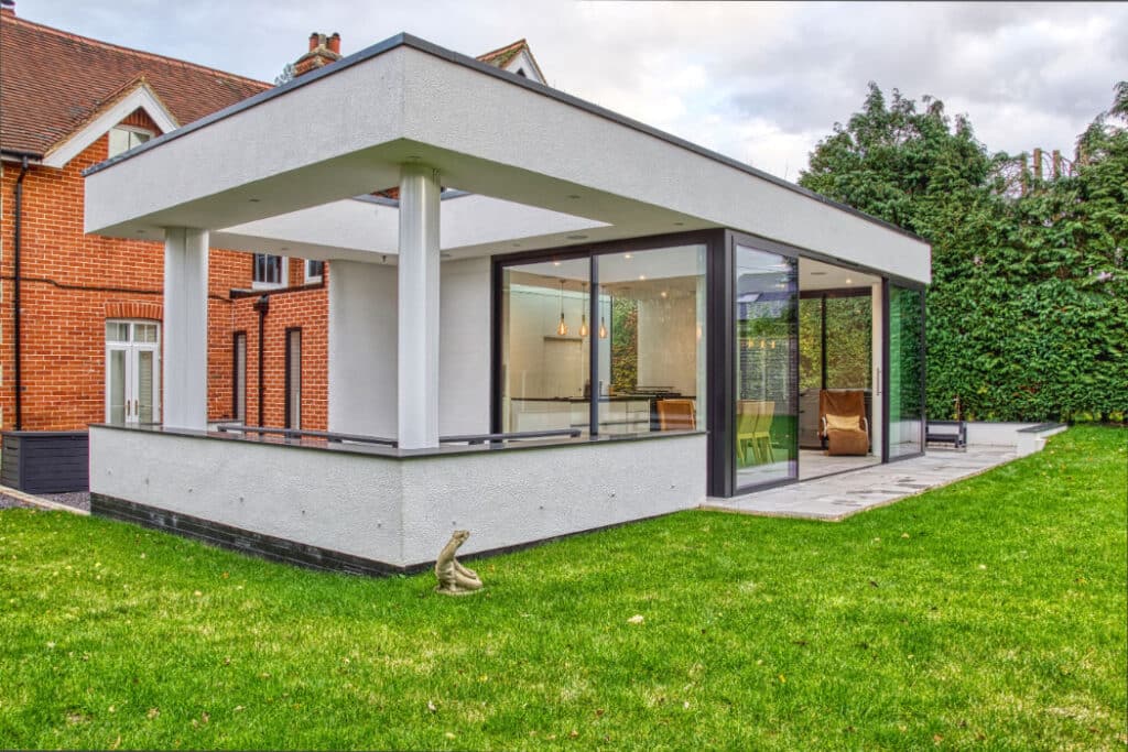 Modern glass and white concrete extension on a traditional red-brick house, with a green lawn and surrounding trees.
