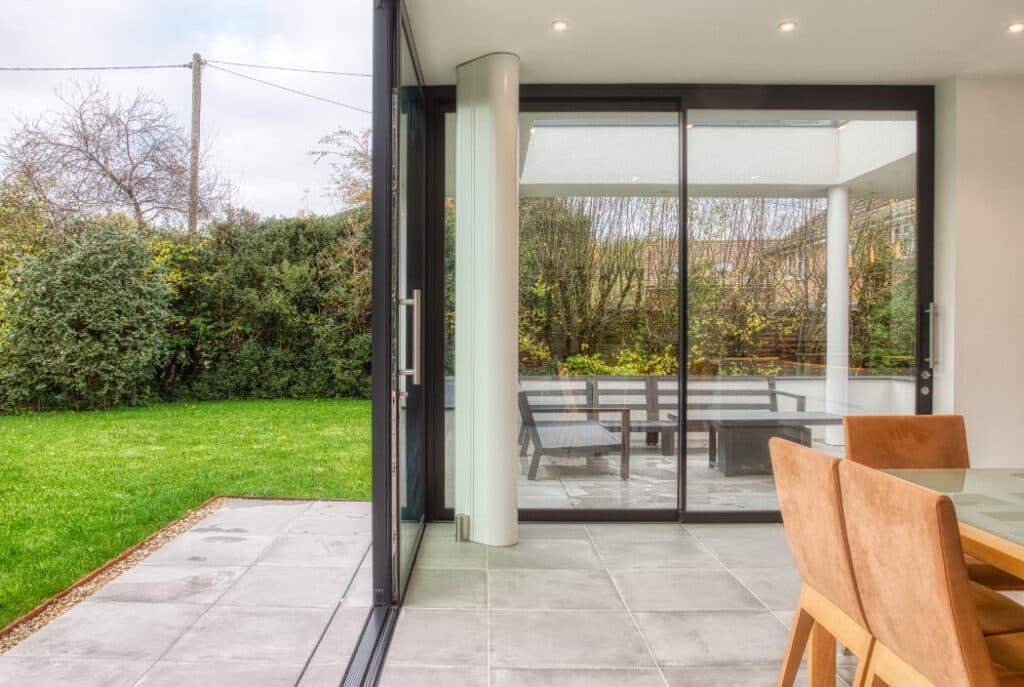 Glass sliding doors open to a patio with a wooden table and chairs, overlooking a green lawn and hedge. Interior features a glass dining table and tan chairs.