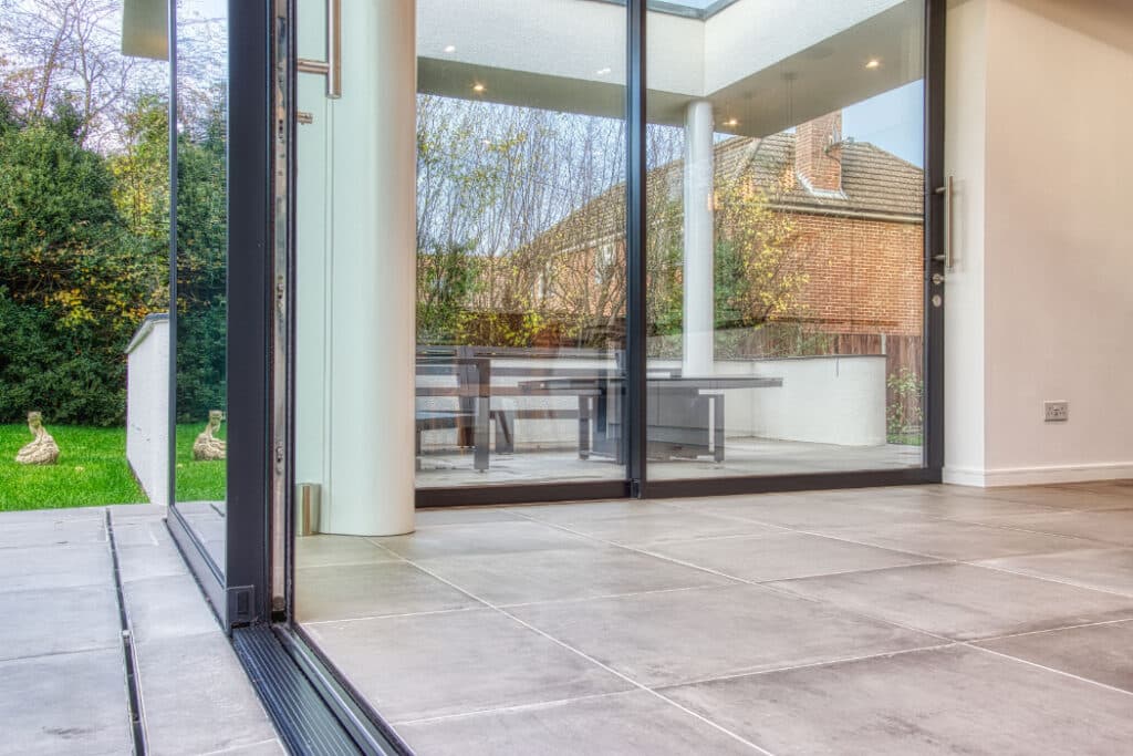 Spacious modern room with large sliding glass doors leading to a patio. The outdoor area includes a wooden table and benches, and glimpses of greenery and brick buildings are visible outside.
