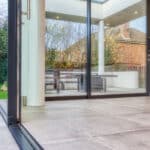 Thumbnail of http://Spacious%20modern%20room%20with%20large%20sliding%20glass%20doors%20leading%20to%20a%20patio.%20The%20outdoor%20area%20includes%20a%20wooden%20table%20and%20benches,%20and%20glimpses%20of%20greenery%20and%20brick%20buildings%20are%20visible%20outside.