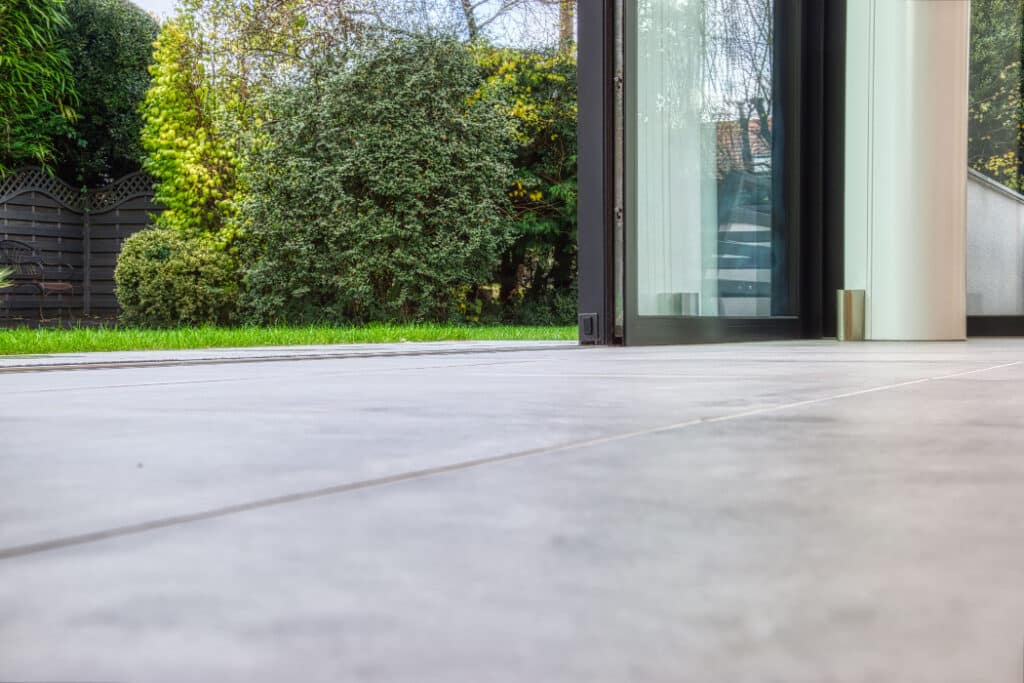 View from a tiled floor through an open glass door to a garden with green grass, bushes, and a wooden fence.