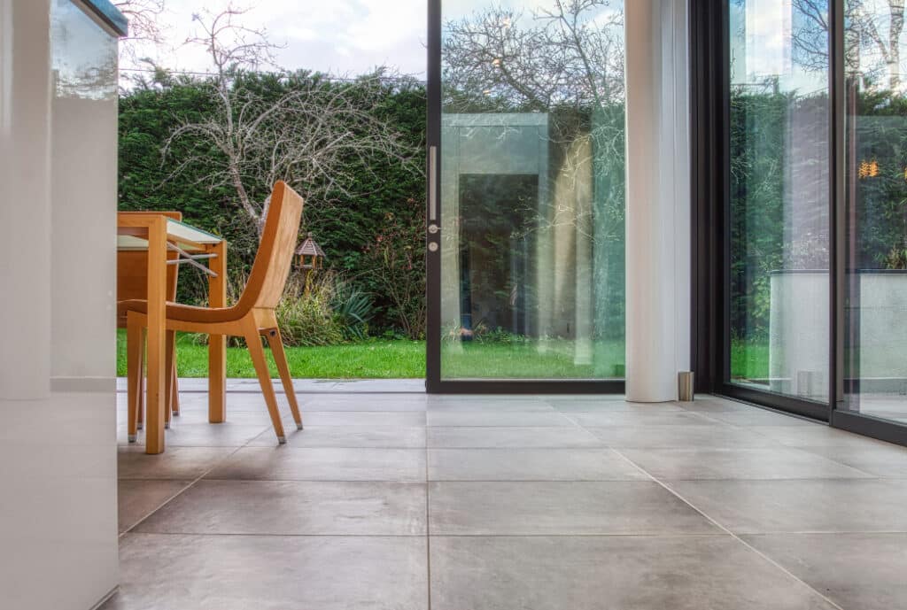 Modern room with large glass sliding doors leading to a green garden. A wooden chair and table are partially visible on the left. The floor is tiled in a neutral color.