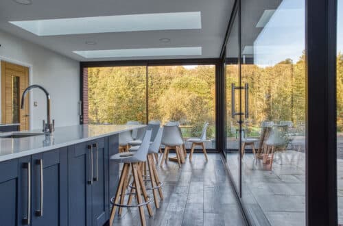 Modern kitchen with a large island, bar stools, and floor-to-ceiling windows showcasing a view of trees. Skylights enhance natural lighting.