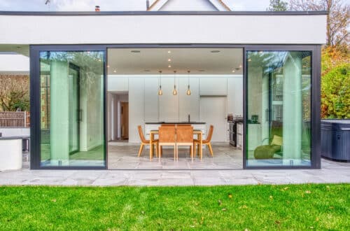Modern kitchen and dining area with large sliding glass doors, wooden table and chairs, and pendant lights, viewed from a green lawn outside.