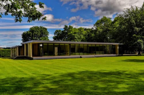 Modern, single-story glass building surrounded by a large green lawn and trees under a partly cloudy sky.