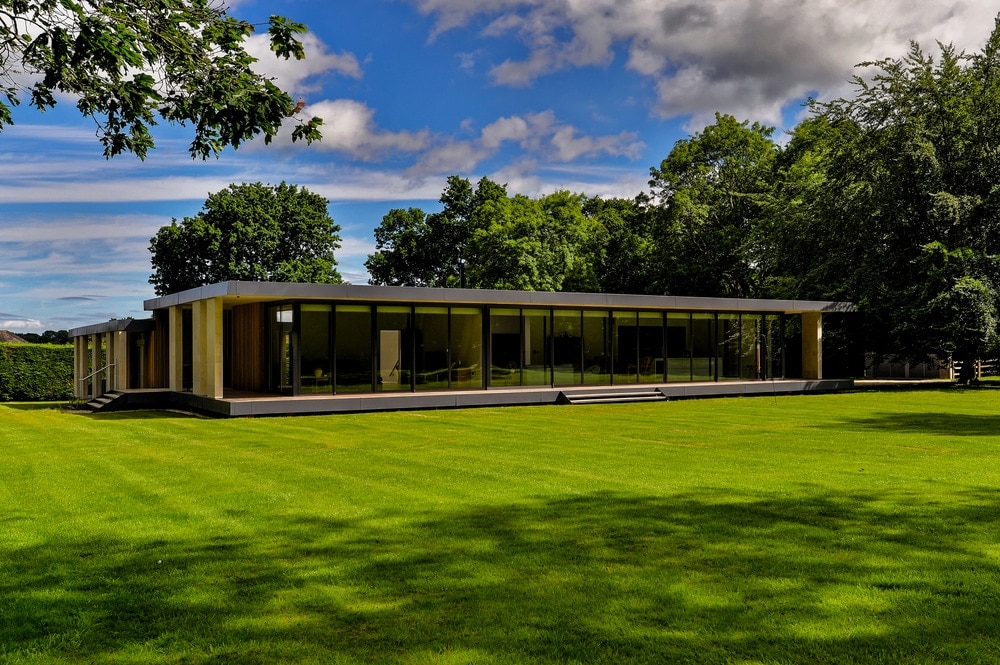 Modern, single-story glass building surrounded by a large green lawn and trees under a partly cloudy sky.