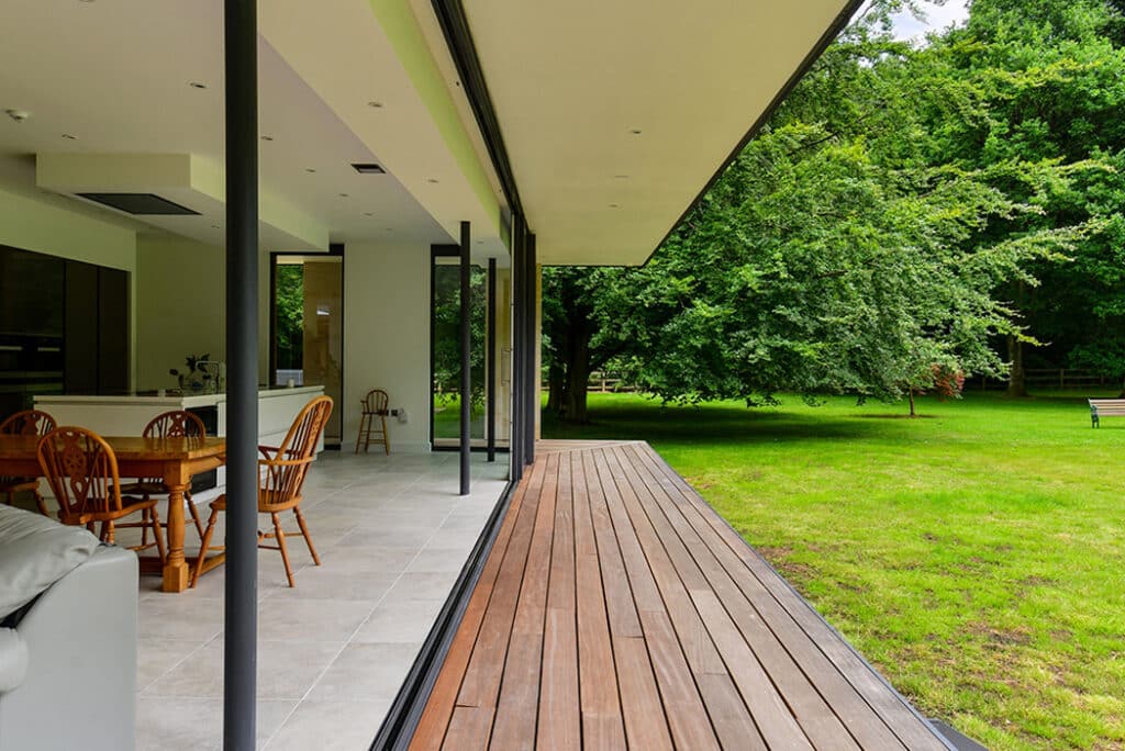 Open modern living space with sliding glass doors leading to a grassy garden. Interior has a wooden dining set on a tiled floor, and an exterior wooden deck is visible.