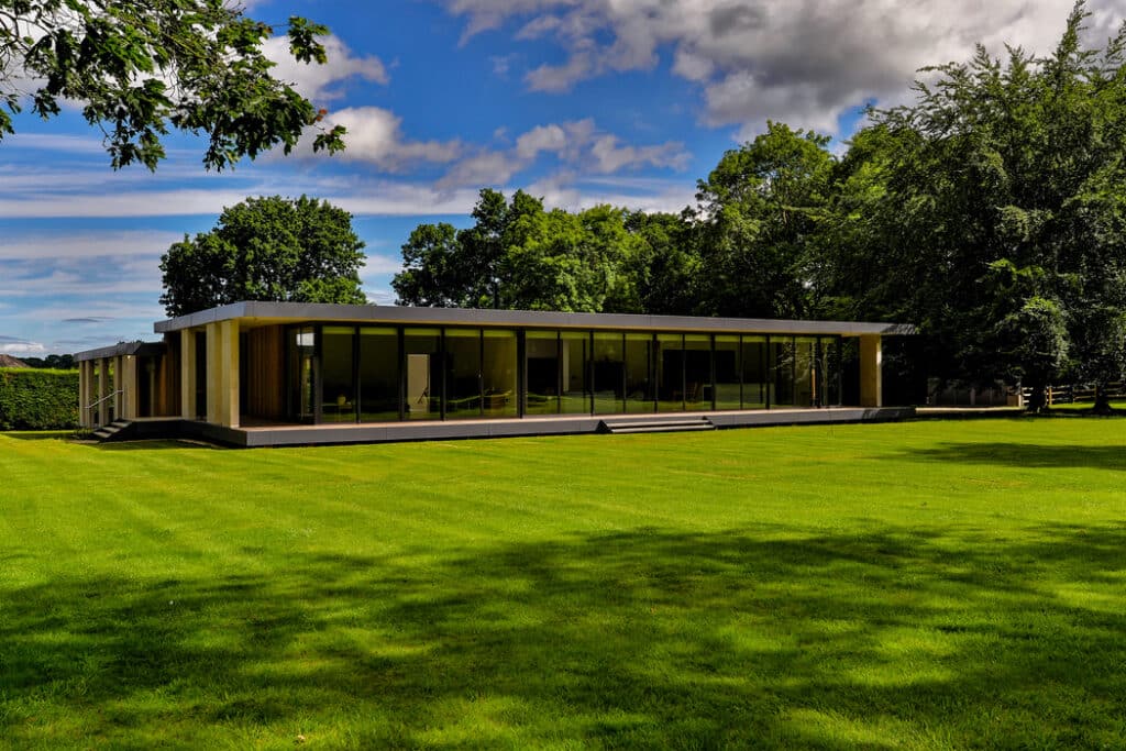 Modern, single-story glass and wood building surrounded by a large, lush green lawn and trees under a partly cloudy sky.