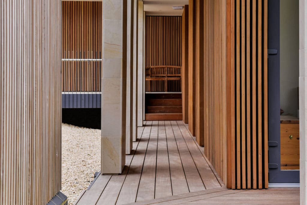 Wooden hallway with vertical slat walls and a matching floor. Staircase at the end leading to a wooden chair. Light filters in from the open side.