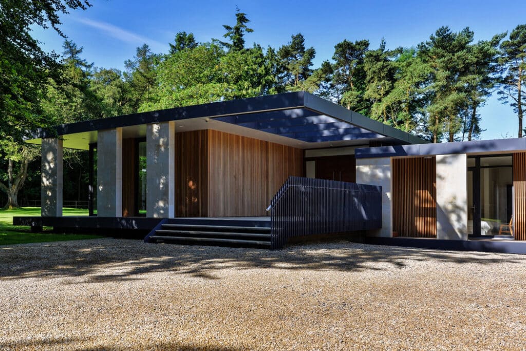 Modern house with flat roof and large windows, surrounded by trees. The building features wood accents and a gravel driveway.