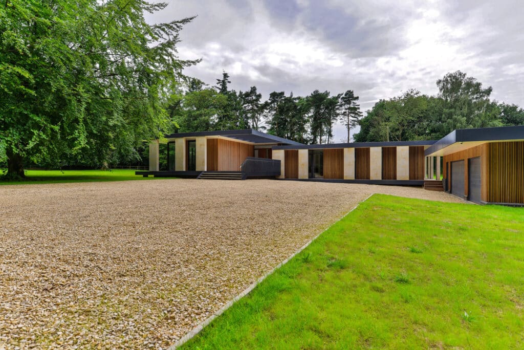 Modern single-story house with large windows, surrounded by trees and a gravel driveway, next to a well-maintained lawn.
