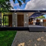 Thumbnail of http://Modern%20single-story%20house%20with%20large%20glass%20windows,%20flat%20roof,%20and%20wooden%20accents.%20Surrounded%20by%20greenery,%20gravel%20pathway,%20and%20a%20stepped%20entrance.%20Partly%20cloudy%20sky%20above.