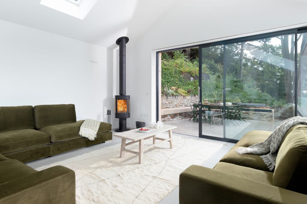 Modern living room with green sofas, a wood-burning stove, and a light rug. Large sliding glass doors open to an outdoor patio with a dining area.