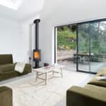 Thumbnail of http://Modern%20living%20room%20with%20green%20sofas,%20a%20wood-burning%20stove,%20and%20a%20light%20rug.%20Large%20sliding%20glass%20doors%20open%20to%20an%20outdoor%20patio%20with%20a%20dining%20area.
