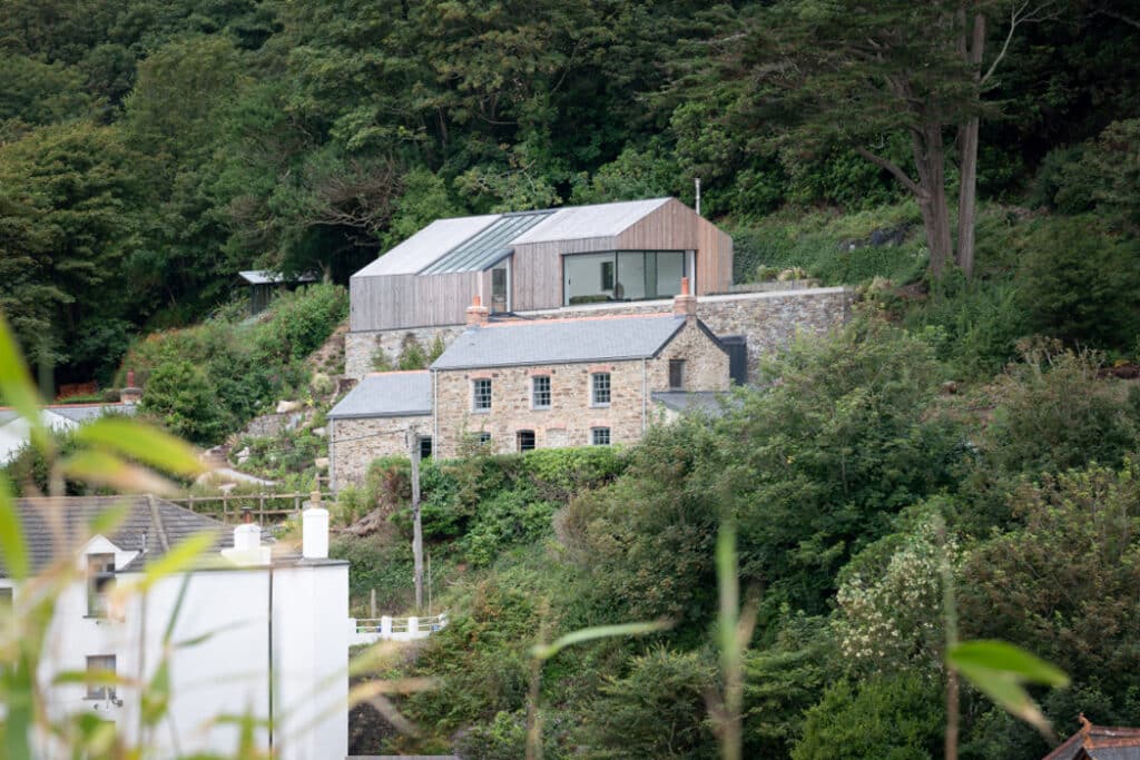 A rustic stone house with a modern wooden extension is nestled in a lush, green hillside. Trees surround the property, and a white building is partially visible in the foreground.