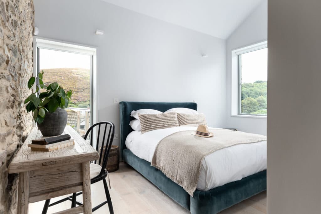 Bright bedroom with a stone wall, featuring a double bed with beige bedding, a wooden desk with a black chair, a potted plant, and two large windows with views of hills and greenery.
