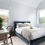 Thumbnail of http://Bright%20bedroom%20with%20a%20stone%20wall,%20featuring%20a%20double%20bed%20with%20beige%20bedding,%20a%20wooden%20desk%20with%20a%20black%20chair,%20a%20potted%20plant,%20and%20two%20large%20windows%20with%20views%20of%20hills%20and%20greenery.