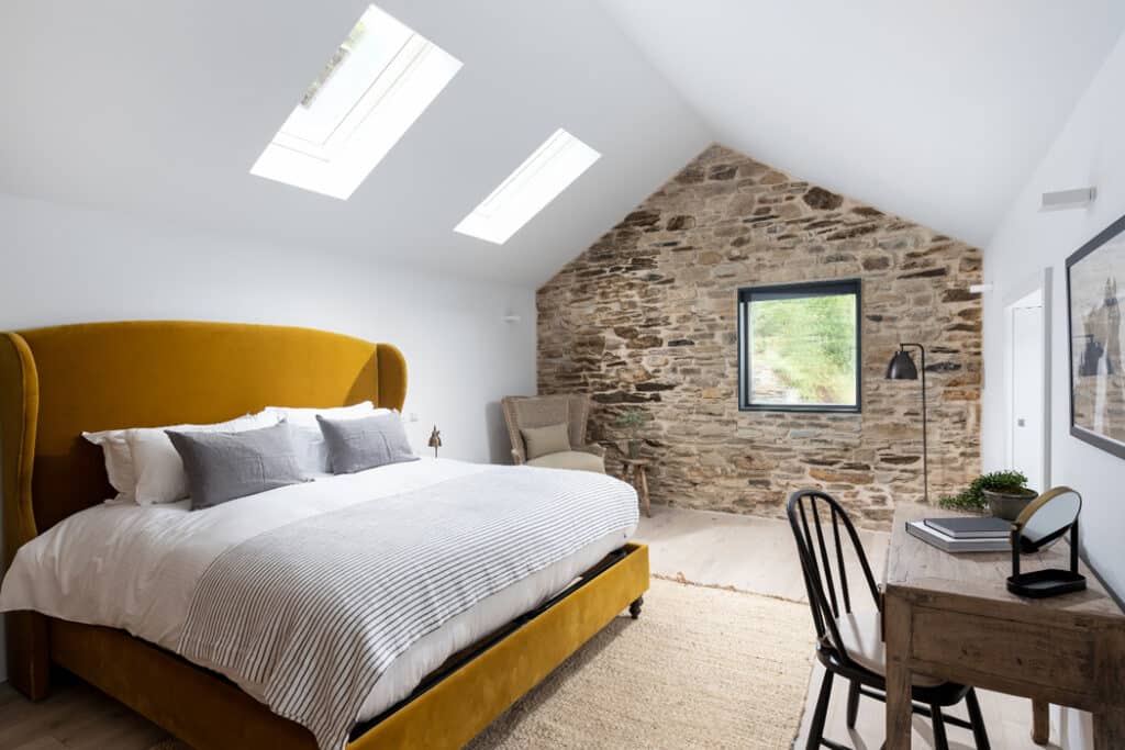Bedroom with a mustard yellow bed, skylights, exposed brick wall, and a window. A desk with a lamp and chair is on the right, and a woven rug is on the floor.