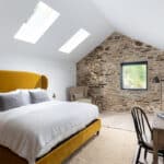 Thumbnail of http://Bedroom%20with%20a%20mustard%20yellow%20bed,%20skylights,%20exposed%20brick%20wall,%20and%20a%20window.%20A%20desk%20with%20a%20lamp%20and%20chair%20is%20on%20the%20right,%20and%20a%20woven%20rug%20is%20on%20the%20floor.