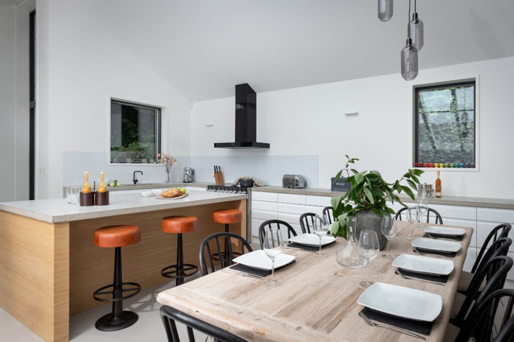 Modern kitchen with a wooden island, three orange stools, and a dining table set for six. White cabinets, black range hood, hanging lights, and two windows enhance natural light.