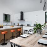 Thumbnail of http://Modern%20kitchen%20with%20a%20wooden%20island,%20three%20orange%20stools,%20and%20a%20dining%20table%20set%20for%20six.%20White%20cabinets,%20black%20range%20hood,%20hanging%20lights,%20and%20two%20windows%20enhance%20natural%20light.