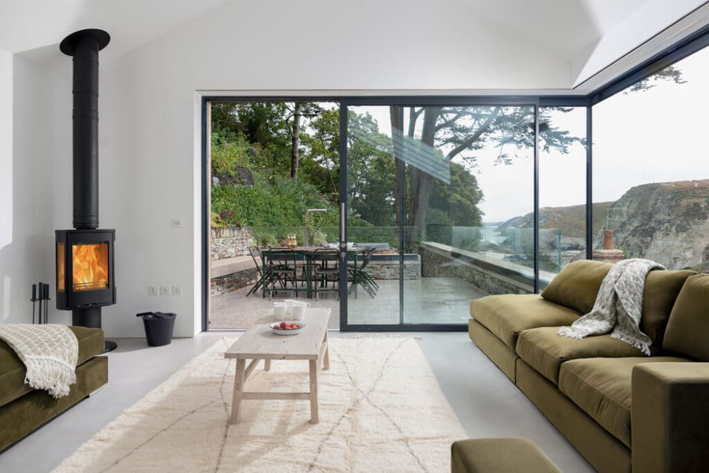 Modern living room with green sofas, a wooden table, and a fireplace. Large glass doors open to a patio with outdoor furniture, overlooking a natural landscape with trees and hills.