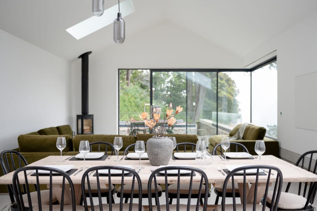 Modern dining area with a wooden table set for eight, black chairs, large windows, green sofas, and a corner fireplace. A vase with orange flowers is centered on the table.