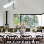 Thumbnail of http://Modern%20dining%20area%20with%20a%20wooden%20table%20set%20for%20eight,%20black%20chairs,%20large%20windows,%20green%20sofas,%20and%20a%20corner%20fireplace.%20A%20vase%20with%20orange%20flowers%20is%20centered%20on%20the%20table.