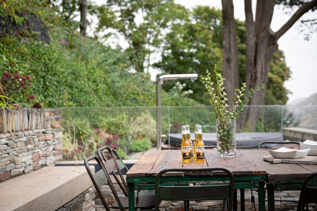 Two bottles of beer on a rustic outdoor table with metal chairs, surrounded by greenery and stone walls, under a cloudy sky.