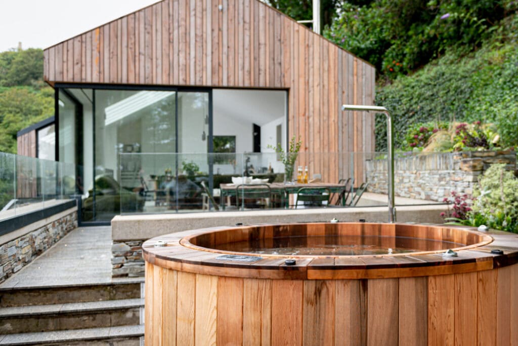 Wooden hot tub in front of a modern glass and wood house, surrounded by plants and stone walls.