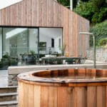 Thumbnail of http://Wooden%20hot%20tub%20in%20front%20of%20a%20modern%20glass%20and%20wood%20house,%20surrounded%20by%20plants%20and%20stone%20walls.