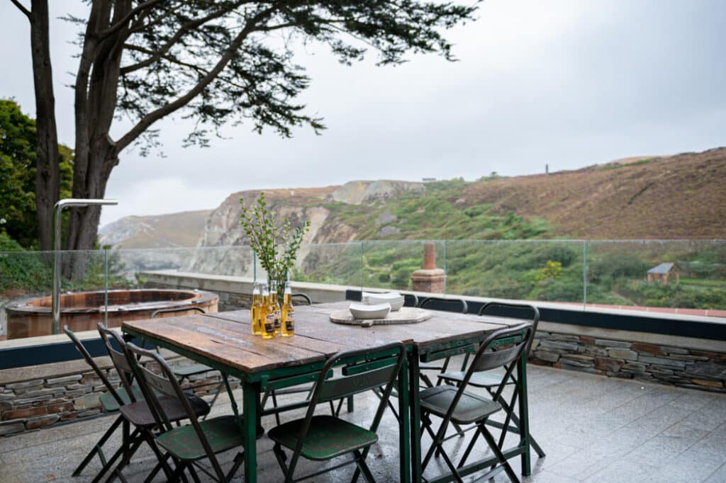 Outdoor dining table with glass bottles and bowls, set on a patio overlooking a hilly landscape with trees and cloudy skies.