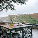 Thumbnail of http://Outdoor%20dining%20table%20with%20glass%20bottles%20and%20bowls,%20set%20on%20a%20patio%20overlooking%20a%20hilly%20landscape%20with%20trees%20and%20cloudy%20skies.