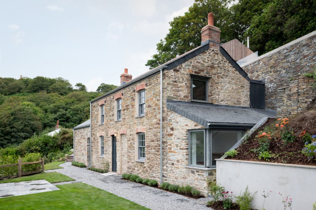 A stone cottage with two chimneys, surrounded by greenery. The building has gray roofing and a path lined with gravel and plants.