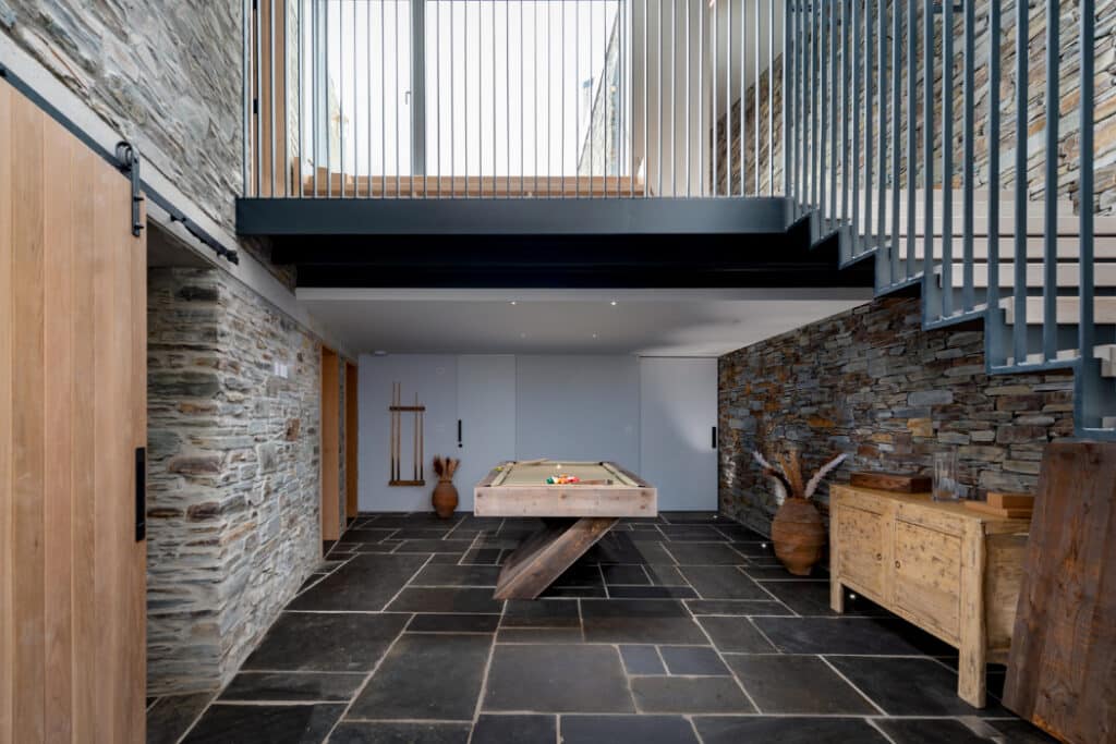 Modern room with stone walls and floor, featuring a pool table in the center. Wooden furniture is placed on the right, and metal stairs lead to an upper level.