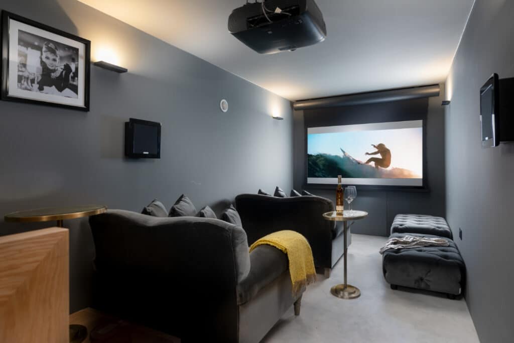 Home theater with gray walls, a ceiling-mounted projector, and a large screen showing a surfer. Black sofas and a yellow blanket in the foreground. Picture on the wall and a small table beside the sofa.