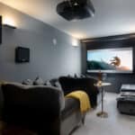 Thumbnail of http://Home%20theater%20with%20gray%20walls,%20a%20ceiling-mounted%20projector,%20and%20a%20large%20screen%20showing%20a%20surfer.%20Black%20sofas%20and%20a%20yellow%20blanket%20in%20the%20foreground.%20Picture%20on%20the%20wall%20and%20a%20small%20table%20beside%20the%20sofa.