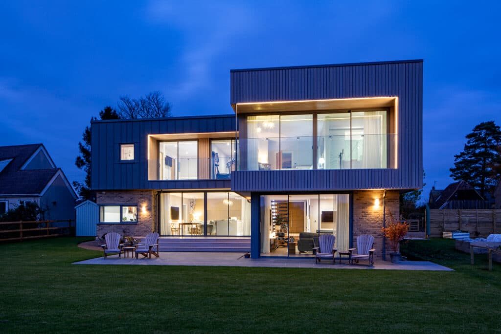 Modern two-story house with large windows and exterior lighting, surrounded by a lawn, deck chairs on a patio, and located in a suburban area at dusk.