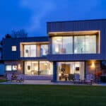 Thumbnail of http://Modern%20two-story%20house%20with%20large%20windows%20and%20exterior%20lighting,%20surrounded%20by%20a%20lawn,%20deck%20chairs%20on%20a%20patio,%20and%20located%20in%20a%20suburban%20area%20at%20dusk.