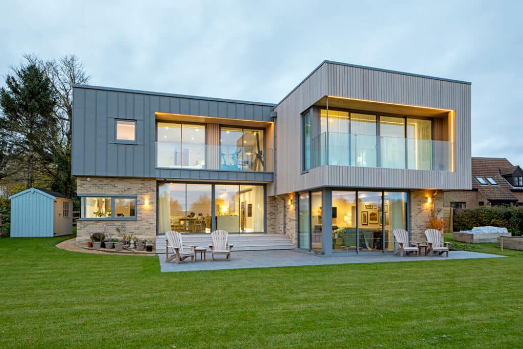 Modern two-story house with large windows, wooden accents, and a well-kept lawn. There are patio chairs on a stone-paved area and a small shed in the background.