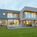 Thumbnail of http://Modern%20two-story%20house%20with%20large%20windows,%20wooden%20accents,%20and%20a%20well-kept%20lawn.%20There%20are%20patio%20chairs%20on%20a%20stone-paved%20area%20and%20a%20small%20shed%20in%20the%20background.