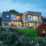 Thumbnail of http://Modern%20two-story%20house%20with%20large%20windows,%20surrounded%20by%20a%20garden.%20A%20round%20metal%20sculpture%20is%20in%20the%20foreground,%20and%20a%20neighboring%20house%20is%20visible%20in%20the%20background.