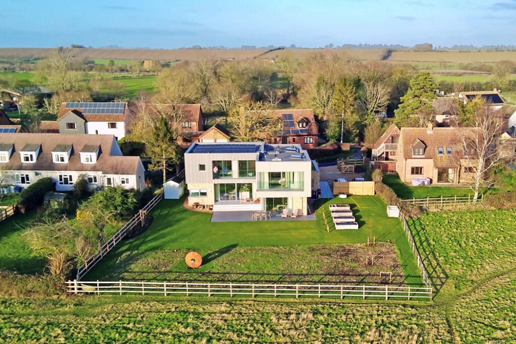 A large modern house with a glass facade is situated in a rural area, surrounded by traditional homes, green fields, and trees.